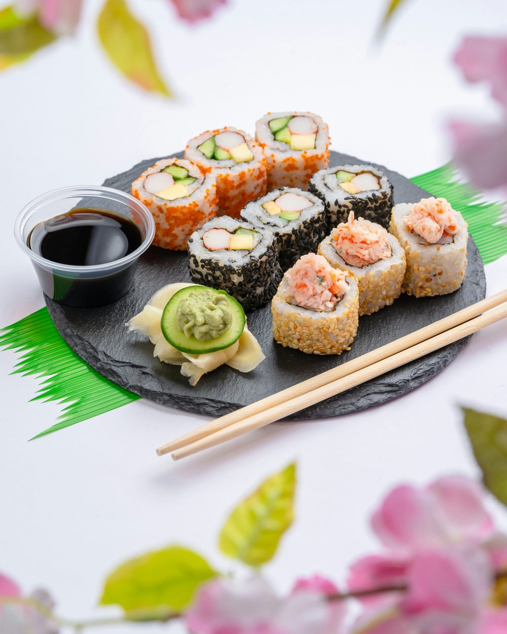 A slate plate of sushi rolls with soy sauce, wasabi, and chopsticks, framed by cherry blossoms