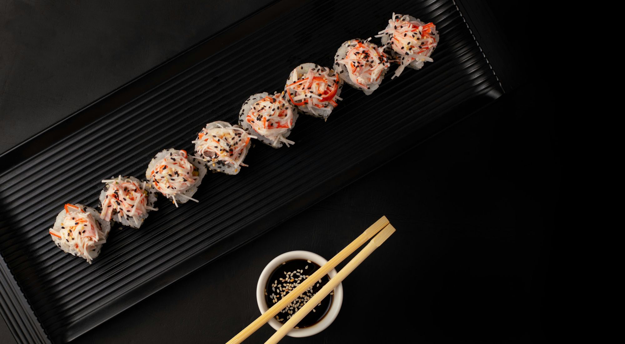 Crab-topped sushi rolls on a long black plate, with soy sauce and chopsticks